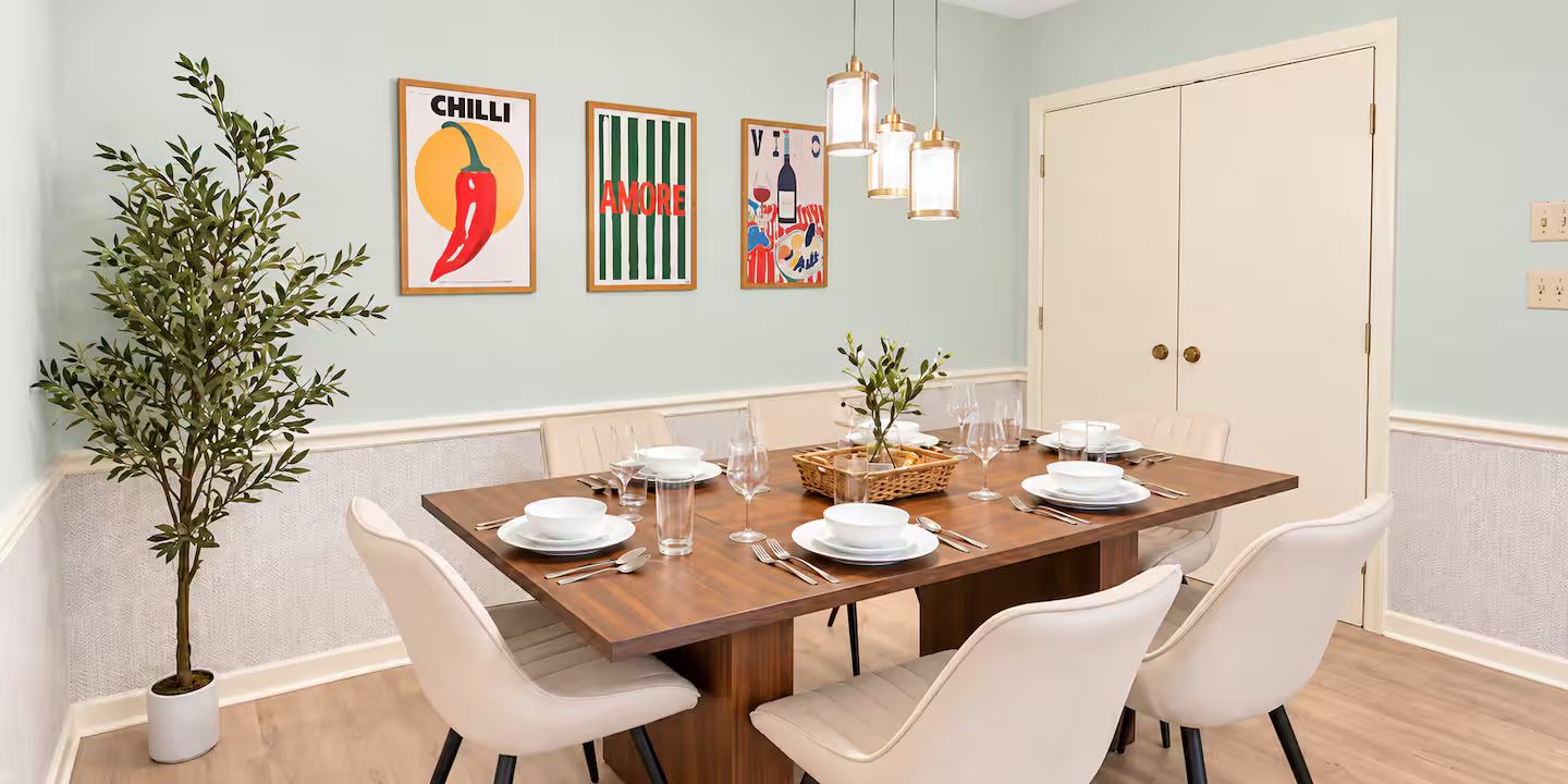 Un coin repas avec une table en bois dressée pour quatre personnes, entourée de chaises blanches, avec une plante en pot sur le côté gauche et une œuvre d’art accrochée au mur au-dessus.