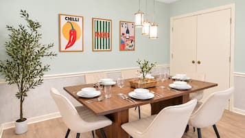 Un coin repas avec une table en bois dressée pour quatre personnes, entourée de chaises blanches, avec une plante en pot sur le côté gauche et une œuvre d’art accrochée au mur au-dessus.