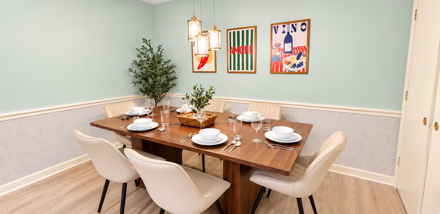 A dining room with a wooden table set for six, white chairs, a potted plant, and wall art.