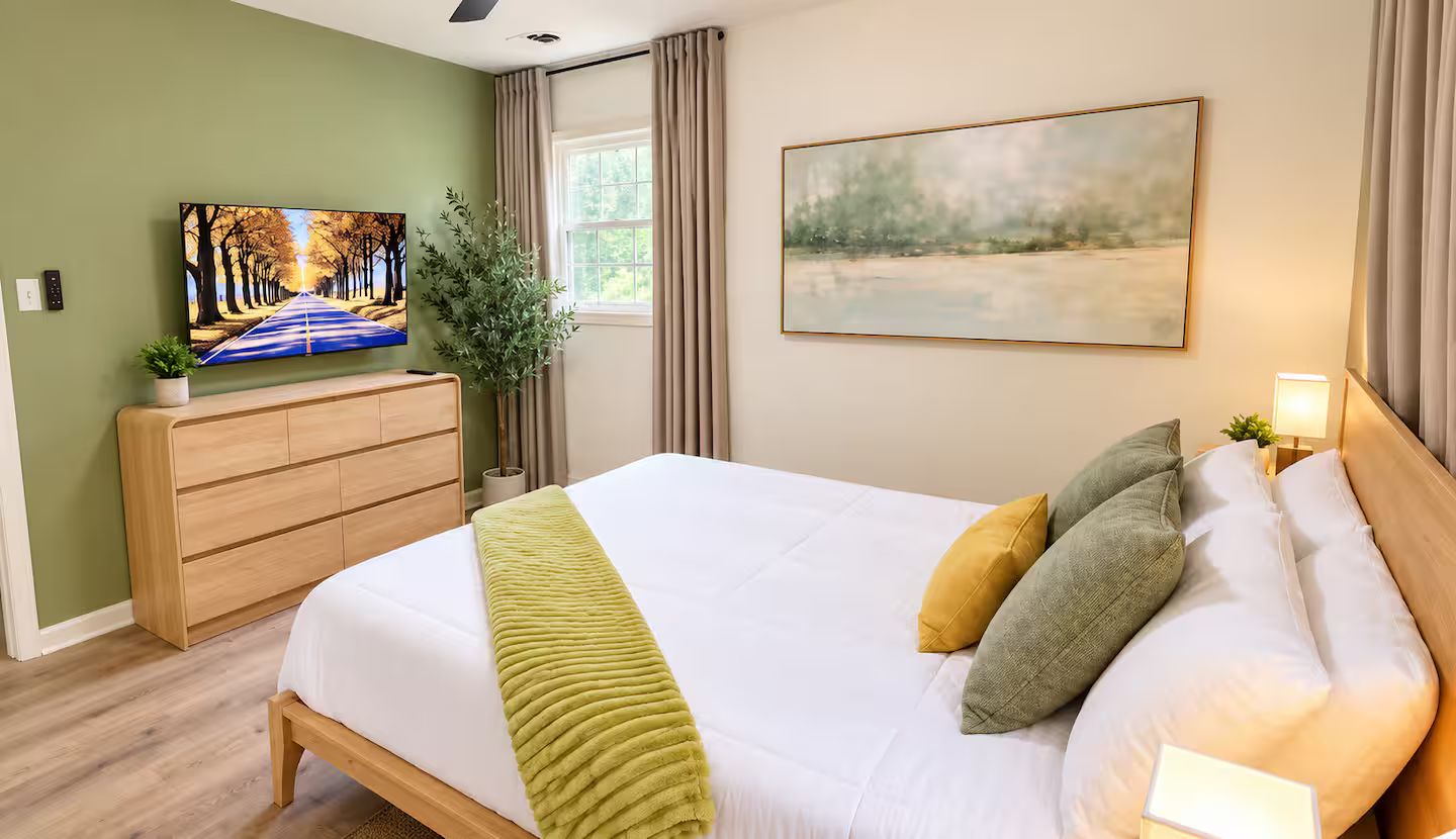 A bedroom with a bed, a television, a window, and a painting on the wall.