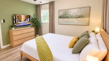 A bedroom with a bed, a television, a window, and a painting on the wall.