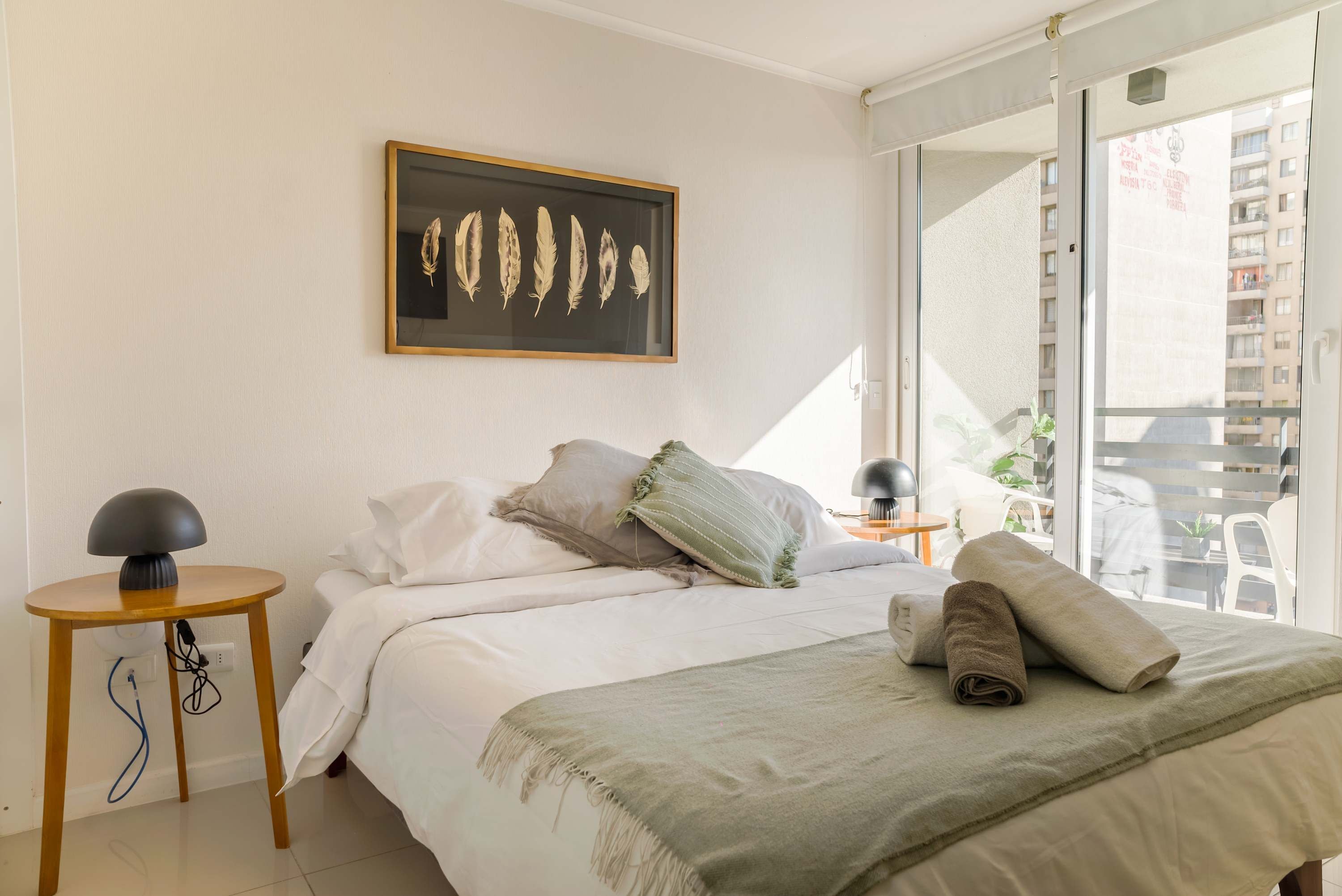 Un dormitorio con cama, almohadas, una mesita de noche con lámpara, una obra de arte enmarcada en la pared y vistas al exterior.