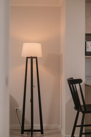 A floor lamp with a white shade, a black chair, and a white wall with a power outlet. - Santa Rosa Apartments by Andes (Santiago)