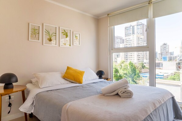 A hotel room with a bed, a bedside table with a lamp, a window with a view of buildings, and framed artwork on the wall. - Santa Rosa Apartments by Andes (Santiago)