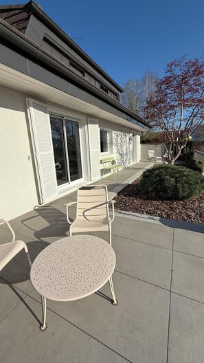 Un bâtiment doté d’un patio comprenant une table ronde et deux chaises, un banc vert et un petit arbre.