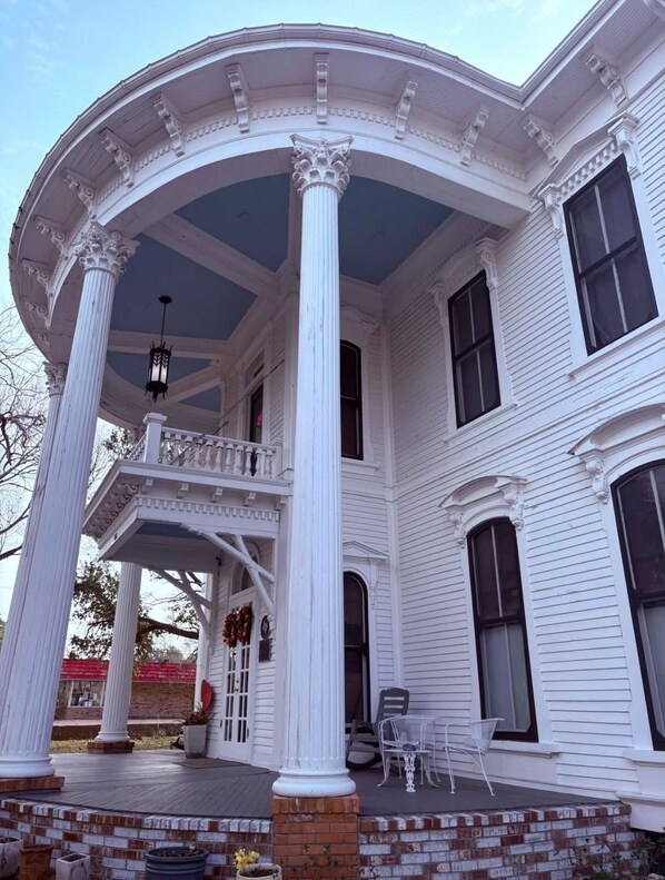 A large, white building with a covered porch supported by columns, featuring a balcony and multiple windows.