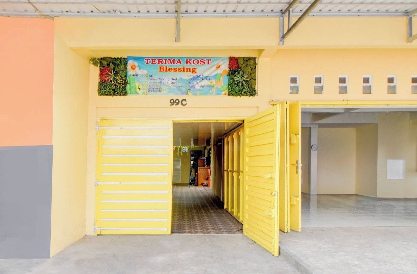 A building entrance with a yellow gate, a sign above reading 'TERIMA KOST Blessing,' and a number '99C' on the wall.