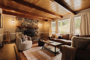 A cozy living room with a stone fireplace, wooden ceiling beams, a sofa, and a coffee table. - Deer & Pine - Hot tub, Dogs ok, Large Patio (Big Bear Lake)