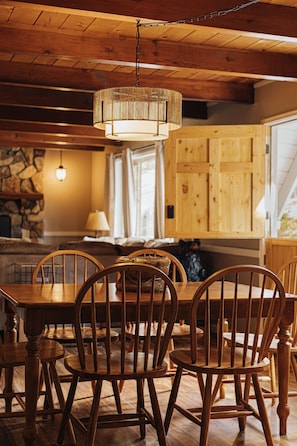 A wooden dining table with chairs, a hanging light fixture, and a stone wall in the background. - Deer & Pine - Hot tub, Dogs ok, Large Patio (Big Bear Lake)