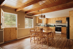 A wooden interior with a dining table and chairs, a kitchen with wooden cabinets, and a large window providing natural light. - Deer & Pine - Hot tub, Dogs ok, Large Patio (Big Bear Lake)