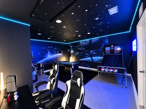Een speelkamer met racezitjes, een game bureau, een basketbalring en een muurschildering met een ruimte thema.