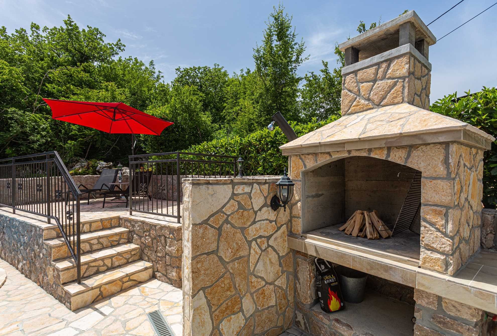 A stone-built outdoor kitchen with a chimney, a red umbrella, and a stone staircase leading to a patio area.