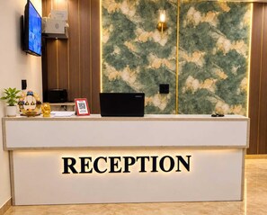 A reception desk with a 'RECEPTION' sign, a flat-screen TV, a Lenovo laptop, a small plant, and a decorative item. - Classic Charmwood Inn (Faridabad)