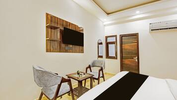 A hotel room with a bed, two chairs, a small table, a flat-screen TV mounted on a wooden panel, a door, and an air conditioning unit.