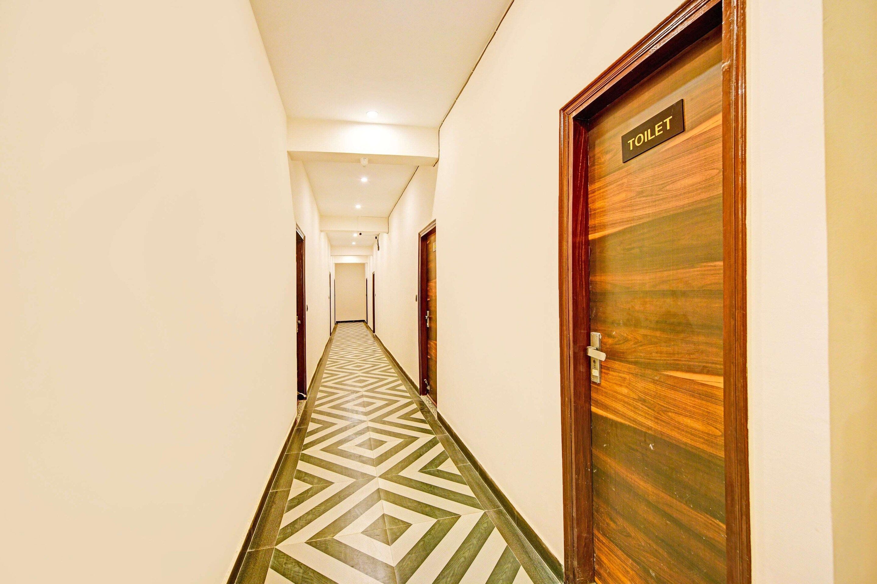 A hotel corridor with a geometric patterned floor, a wooden door labeled 'TOILET' on the right, and a hallway leading to a room.