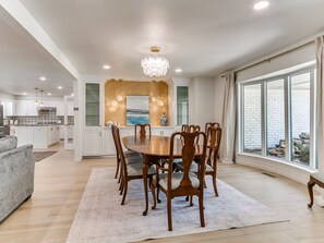 A spacious dining area with a large wooden table and chairs, a chandelier, and a view of a brick exterior through large windows. - Quail Creek Home | Koi Pond, 2 Kings, EV Charger (Oklahoma City)