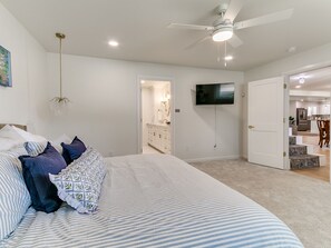 A bedroom with a bed, pillows, a ceiling fan, a TV, and a bathroom visible through an open door. - Quail Creek Home | Koi Pond, 2 Kings, EV Charger (Oklahoma City)