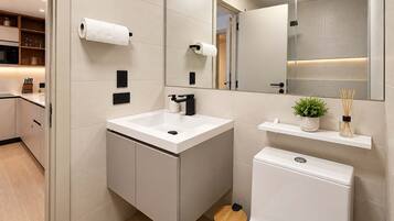 A modern bathroom with a white sink, black faucet, and a toilet. There are two rolls of toilet paper on the wall, a small trash bin, and a shelf with a plant and reed diffuser.