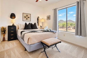 A bedroom with a bed, bedside tables, a dresser, a bench, and a large window with a view of the desert. - Cave Creek Desert Dream — 3BR Main House (Cave Creek)