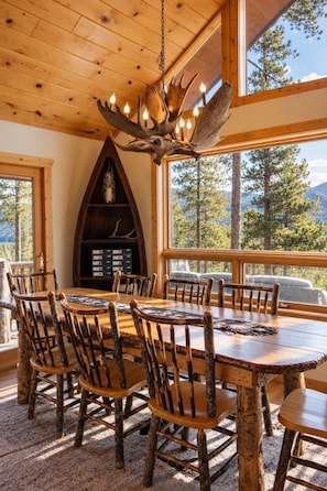 A rustic dining area with a large wooden table and chairs, a chandelier with antlers, and a fireplace with a wooden mantel. - Lakeview Retreat Walk to Town + Fenced Yard (Grand Lake)