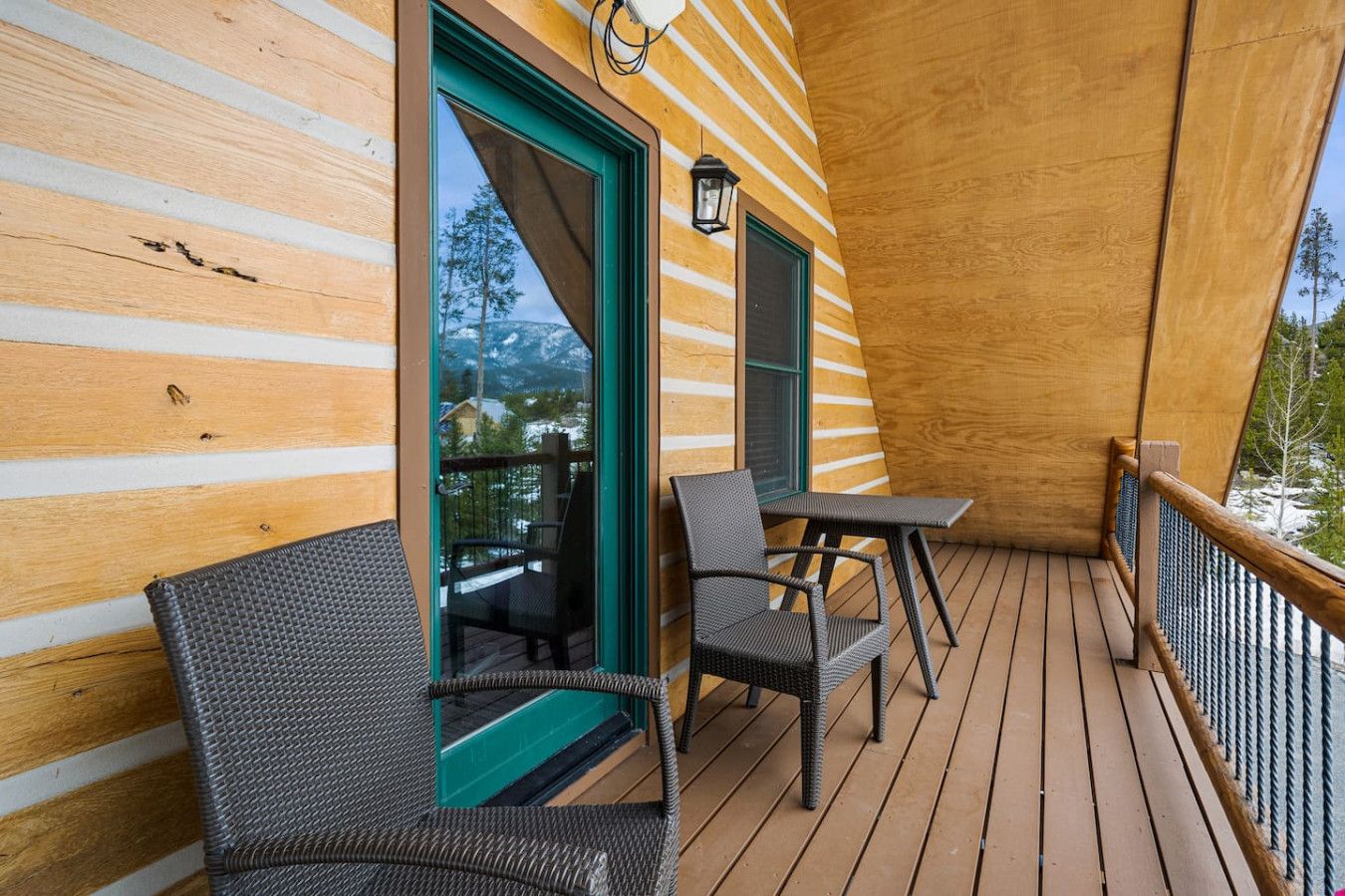Een houten terras met twee rieten stoelen en een kleine tafel, een glazen deur naar buiten en een lantaarn aan de muur.