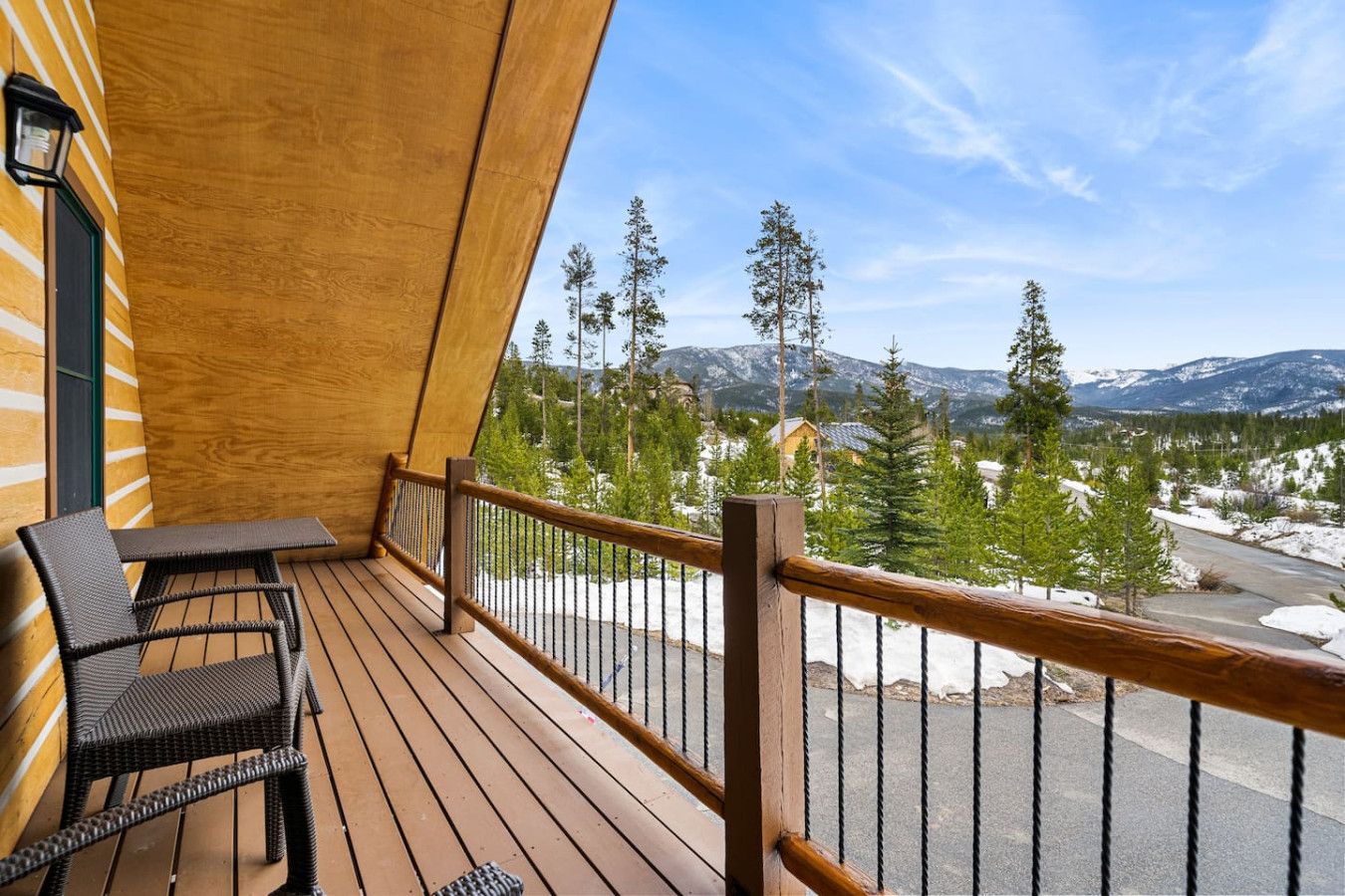 Une terrasse en bois avec une balustrade, deux fauteuils en osier et une vue sur des montagnes et des arbres enneigés.