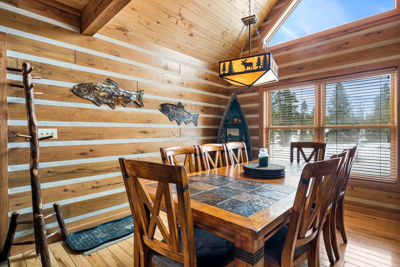 A wooden cabin interior with a dining table, chairs, a fish wall art, a hanging light fixture, and a window with a view of trees.