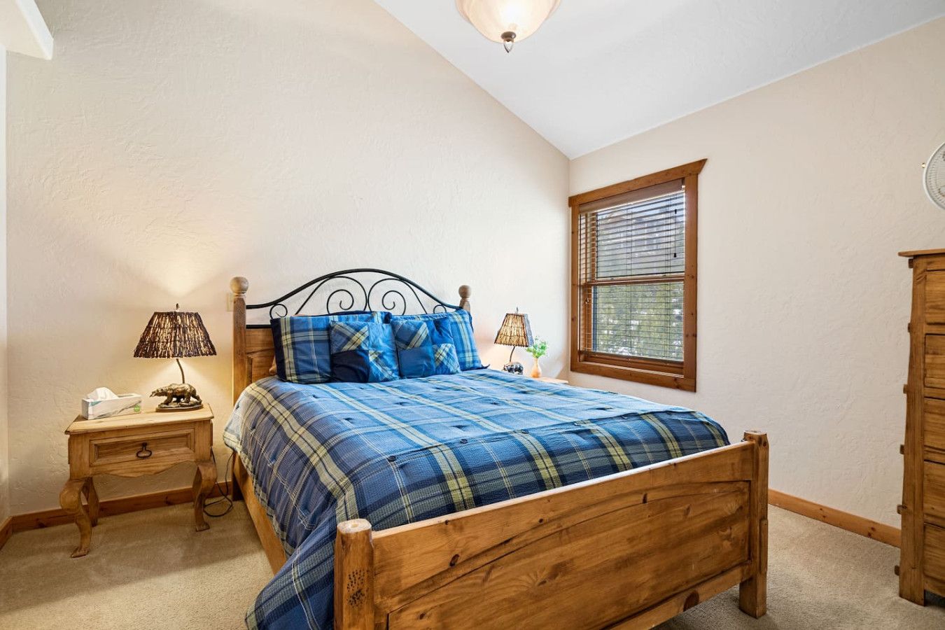 A bedroom with a wooden bed, a nightstand with a lamp, a window with blinds, and a wooden wardrobe.