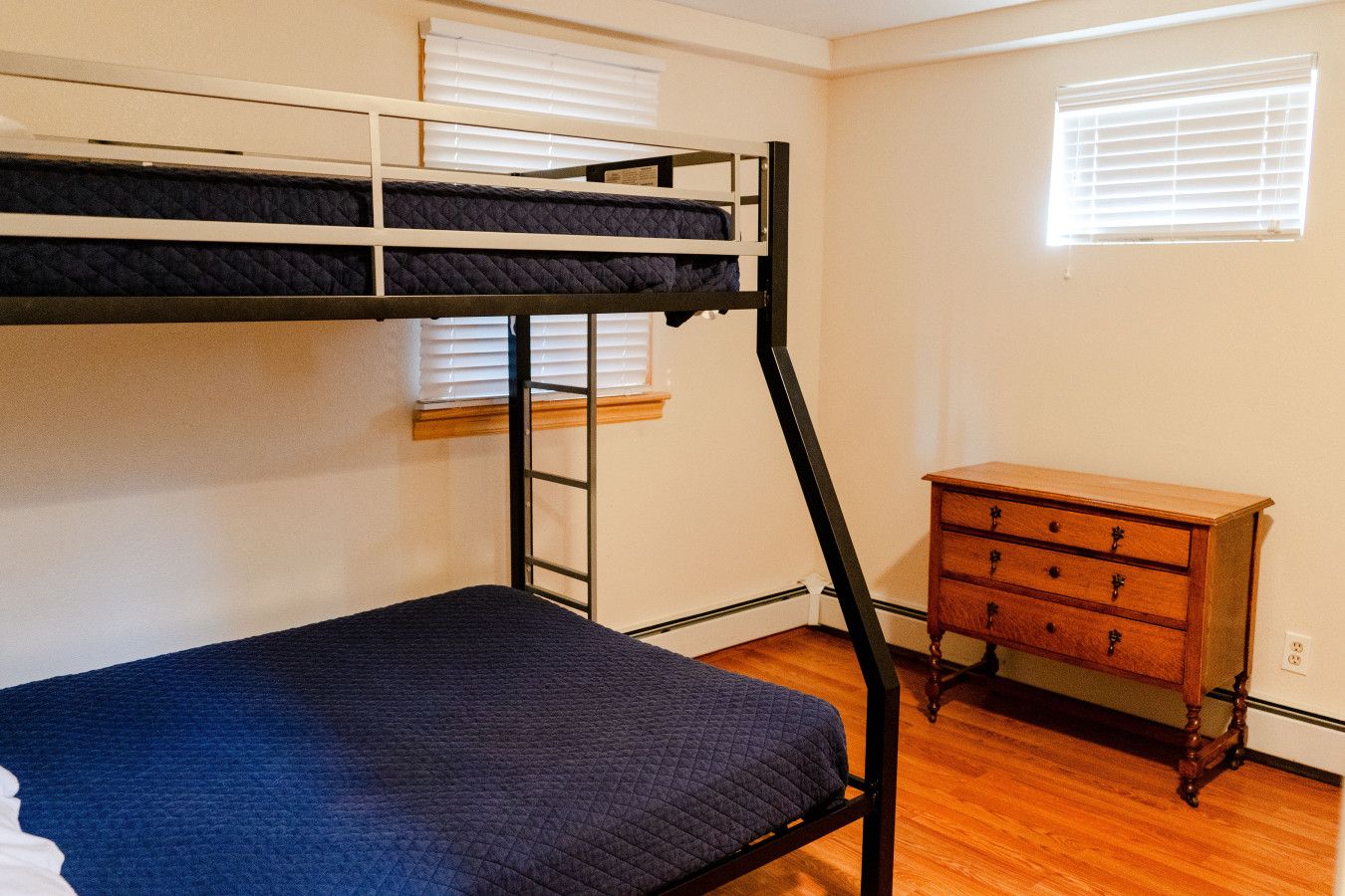 A bunk bed with a wooden dresser and a window with blinds.