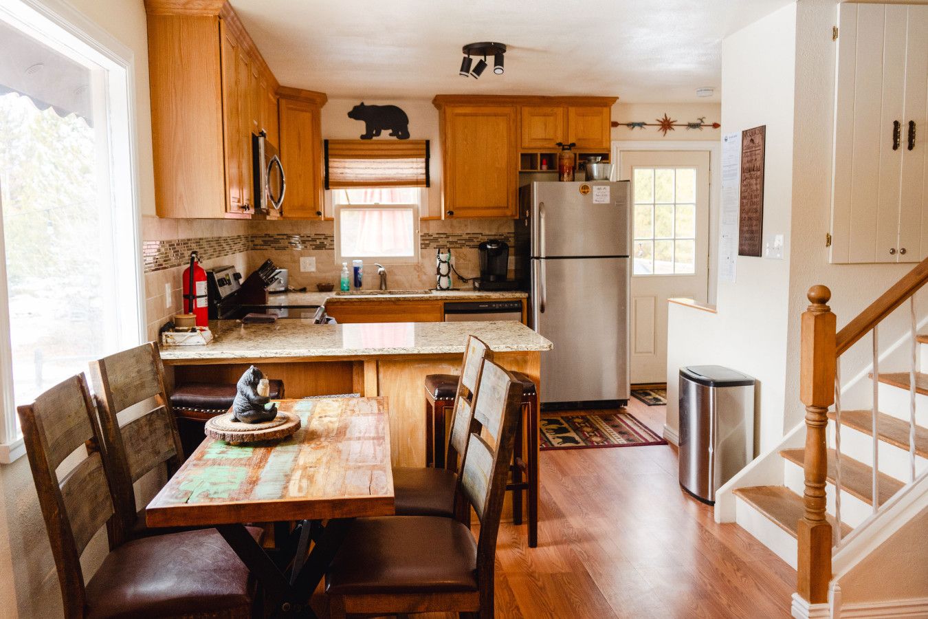 A kitchen with wooden cabinets, a stainless steel refrigerator, a dining table with chairs, and a bear figurine on the table.