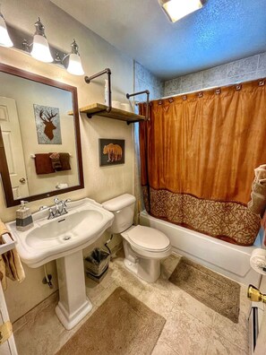 Een badkamer met een witte wastafel, toilet en bad. Er zijn drie wandlampen, een handdoekhouder en een spiegel.