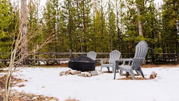 A snow-covered outdoor seating area with Adirondack chairs and a covered grill, surrounded by a wooden fence and tall pine trees.