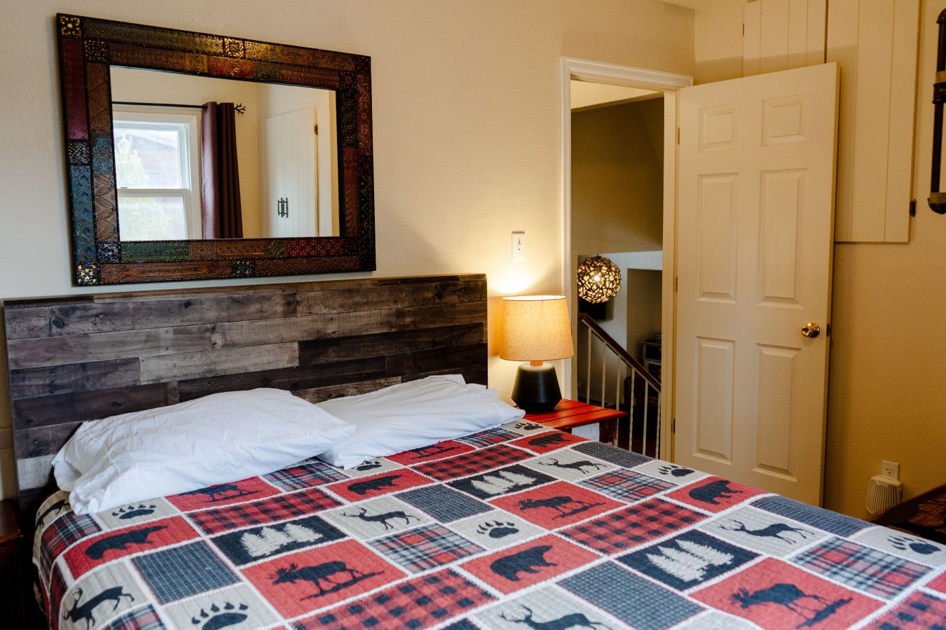 Un dormitorio con cabecera de madera, colcha con estampados de animales, espejo, lámpara y puerta que da a otra habitación.