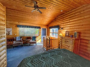 A log cabin bedroom with a bed, two chairs, a small table, a dresser, and a ceiling fan.