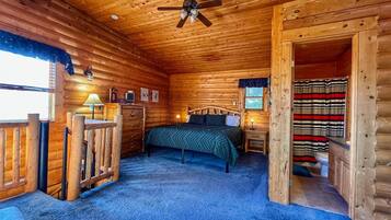 A log cabin bedroom with a bed, wooden walls, a ceiling fan, and a view of the outside.