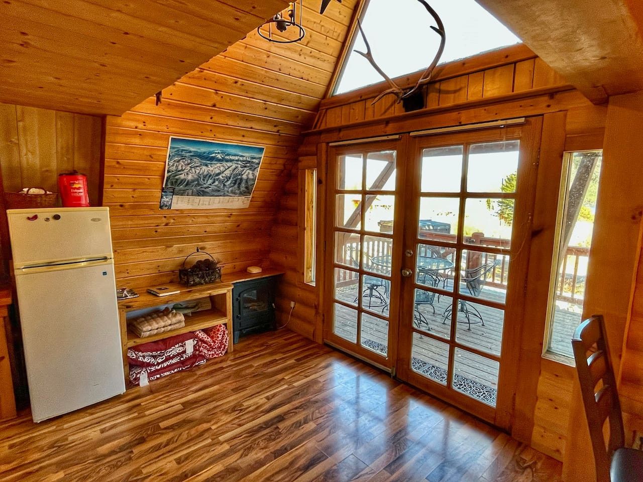 A cozy cabin interior with a wooden ceiling, a refrigerator, a small table, and a large sliding glass door leading to an outdoor deck.