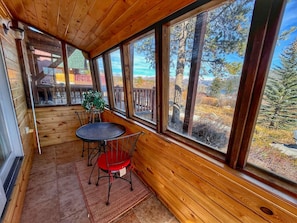 Una cabaña de madera con un porche cubierto, que incluye una mesa y sillas, ventanas amplias y vistas a un paisaje boscoso.