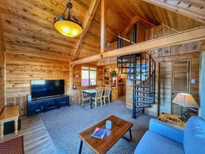 Interior acogedor de una cabaña de troncos, con comedor de madera, televisor de pantalla plana, escalera y sofá.