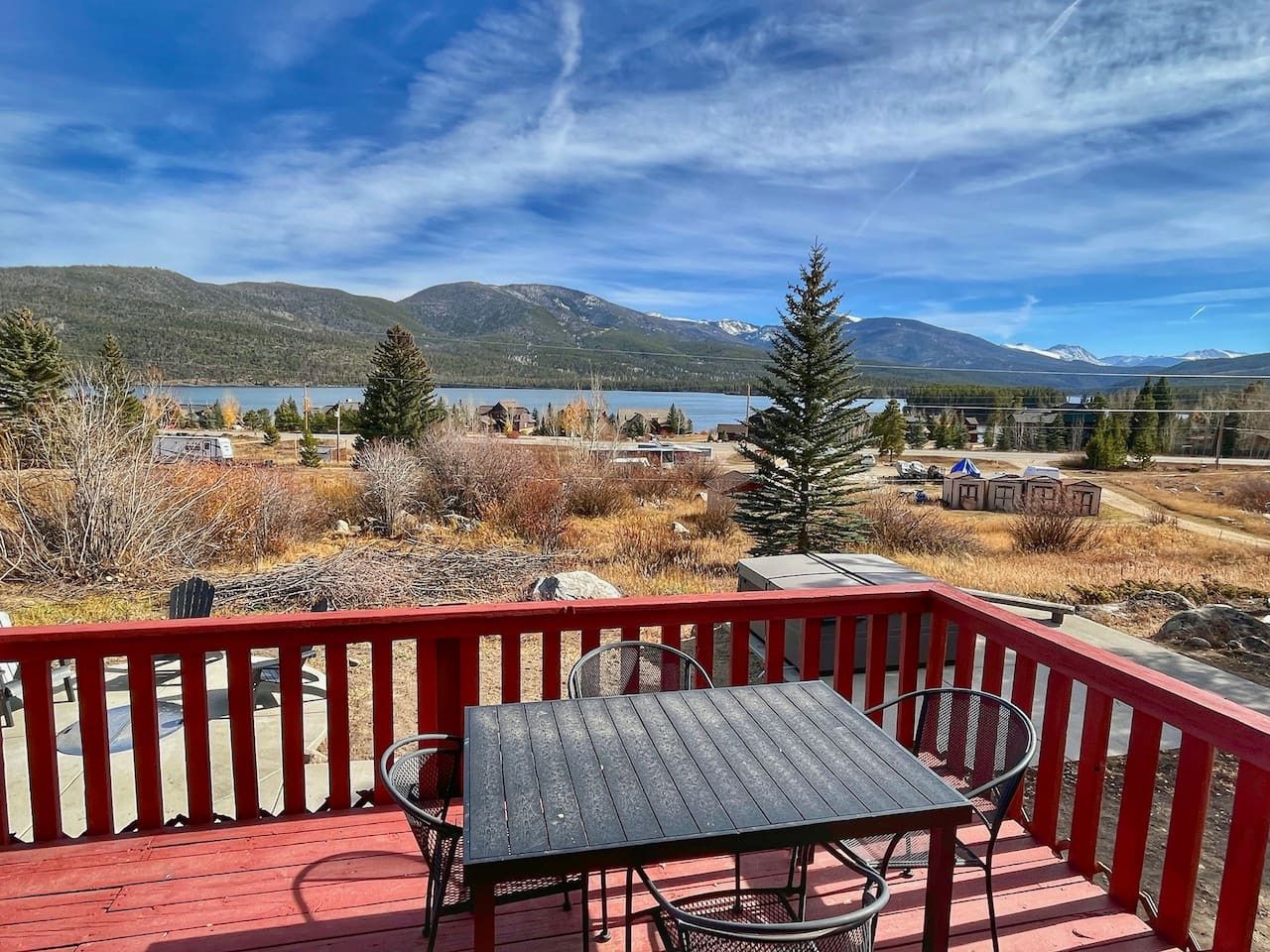 A wooden deck with a table and chairs, overlooking a lake and mountains.