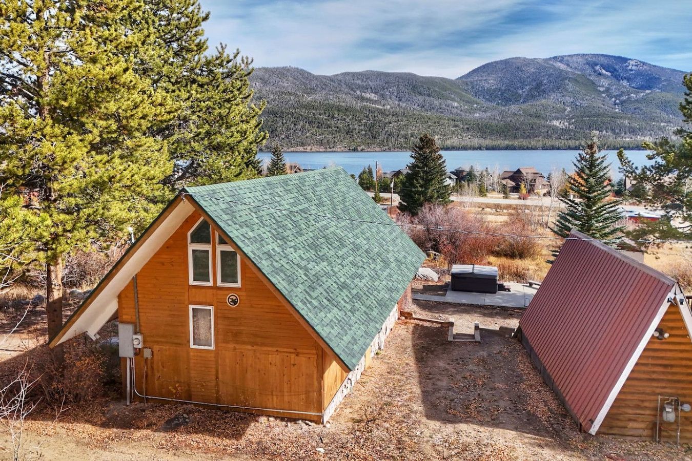 Una cabaña de madera con techo verde, rodeada de árboles y con vistas a un lago y montañas.