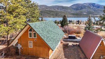 Una cabaña de madera con techo verde, rodeada de árboles y con vistas a un lago y montañas.