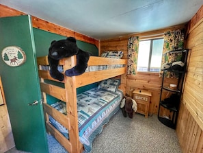 A bunk bed room with a wooden headboard, a small wooden cabinet, and a window with curtains.
