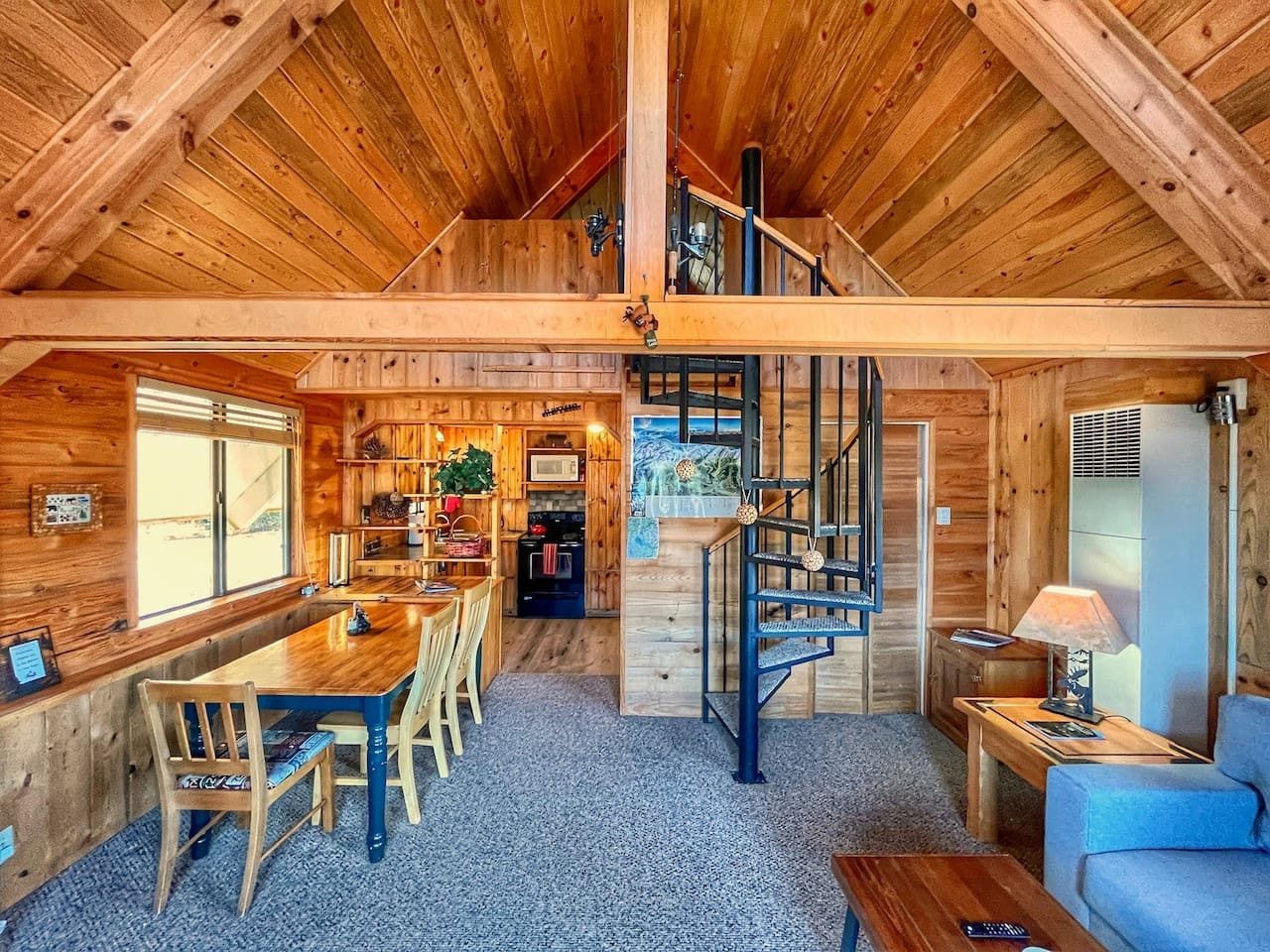 A wooden cabin interior with a dining area, kitchen, and a spiral staircase.