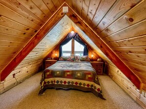 Un dormitorio acogedor en el ático con techo inclinado, una cama grande con colcha estampada, dos mesitas de noche con lámparas y una ventana con cortinas.