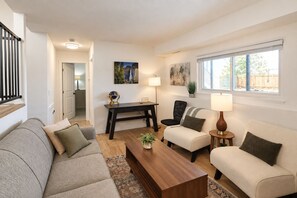 Ein Wohnzimmer mit Sofa, Sesseln, Couchtisch, Schreibtisch und Lampe. Ein Fenster bietet Blick ins Freie.