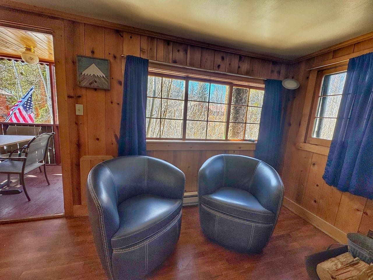 A room with wooden walls and a large window with blue curtains. There are two leather chairs in the center.