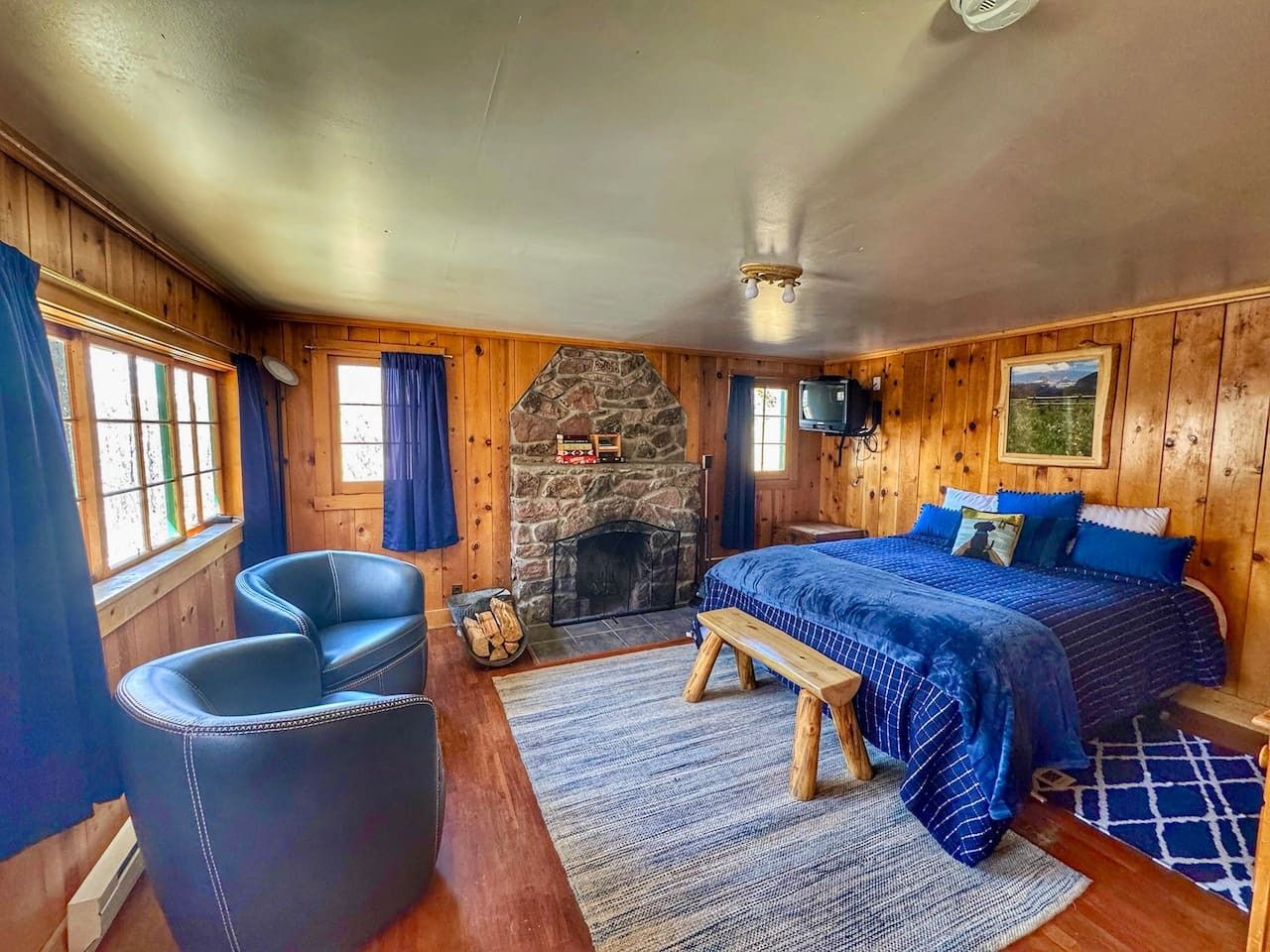 Una habitación acogedora con chimenea de piedra, una cama con ropa de cama azul, un banco y una mesita con un cesto de leña.