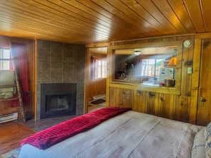 Une pièce chaleureuse avec une cheminée, des murs en bois, un lit et un coin cuisine visible à travers une porte en verre.