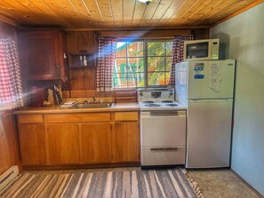 A compact kitchen with wooden cabinets, a stove, a refrigerator, and a window with curtains. - 1 BR with sunroom-walk to beach (Grand Lake)