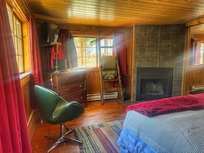A cozy room with a fireplace, wooden walls, a green chair, and a window with curtains. - 1 BR with sunroom-walk to beach (Grand Lake)